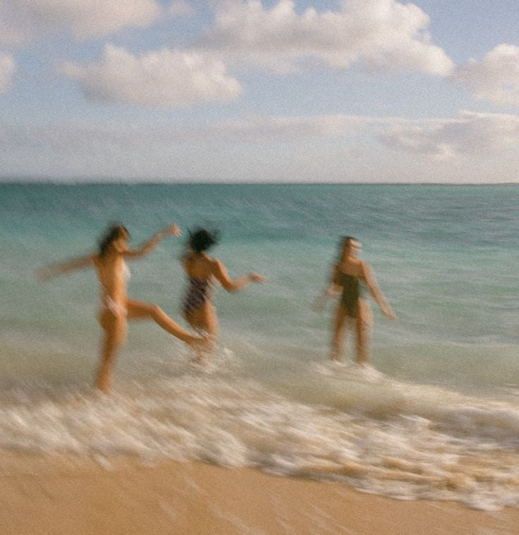 Three people standing in shallow ocean water on a sandy beach under a cloudy sky, captured in a soft, blurry, nostalgic style.